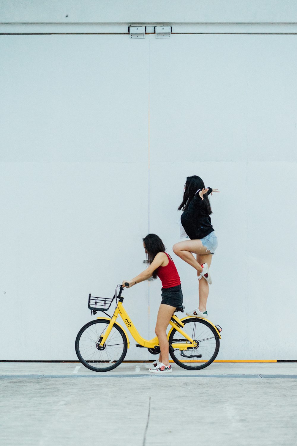 woman riding bicycle with another woman standing on rear fender near wall