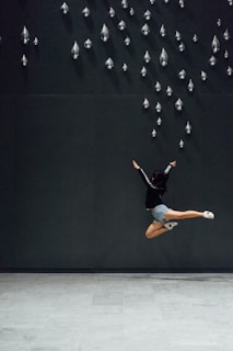 A person is captured in mid-air performing a high jump against a dark wall. They are wearing a black jacket and gray shorts. The wall features numerous metallic, teardrop-shaped sculptures arranged in a pattern.