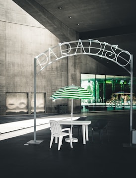 A minimalist outdoor setup with a white table and chairs under a green and white striped umbrella, set beneath a sign with the word 'Paradiso' in neon letters. The setting appears to be in front of a modern building with large glass windows and concrete walls, creating a stark yet stylish contrast.
