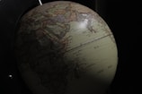 A detailed view of a globe depicting geographic regions, with visible countries and oceans, partially illuminated against a dark background. The continents are labeled, and major cities are marked.