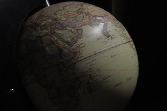 A detailed view of a globe depicting geographic regions, with visible countries and oceans, partially illuminated against a dark background. The continents are labeled, and major cities are marked.