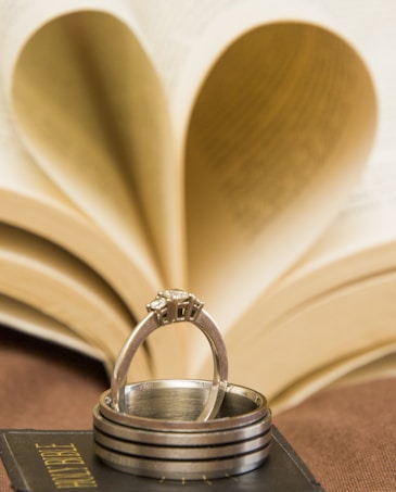 A stack of rings is elegantly placed atop a book, with the background displaying the pages of an open book forming a heart shape. The focus is on the metallic texture of the rings, and the heart-shaped book pages add a romantic element.