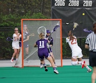 A competitive lacrosse game with female players. One player in a white jersey is attempting to score a goal, while the opposing team in purple, including the goalie, defends the net. The action takes place on a green artificial turf field with a visible scoreboard in the background indicating past championship years.