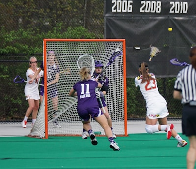 A competitive lacrosse game with female players. One player in a white jersey is attempting to score a goal, while the opposing team in purple, including the goalie, defends the net. The action takes place on a green artificial turf field with a visible scoreboard in the background indicating past championship years.