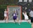 A competitive lacrosse game with female players. One player in a white jersey is attempting to score a goal, while the opposing team in purple, including the goalie, defends the net. The action takes place on a green artificial turf field with a visible scoreboard in the background indicating past championship years.