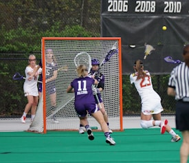 A competitive lacrosse game with female players. One player in a white jersey is attempting to score a goal, while the opposing team in purple, including the goalie, defends the net. The action takes place on a green artificial turf field with a visible scoreboard in the background indicating past championship years.