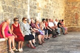 A group of elderly people are seated along a stone wall in a historic setting. They are dressed in casual summer clothing and appear to be engaged in conversation. The wall is made of rustic stone bricks, contributing to an old-world charm.