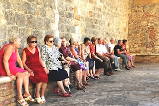 A group of elderly people are seated along a stone wall in a historic setting. They are dressed in casual summer clothing and appear to be engaged in conversation. The wall is made of rustic stone bricks, contributing to an old-world charm.