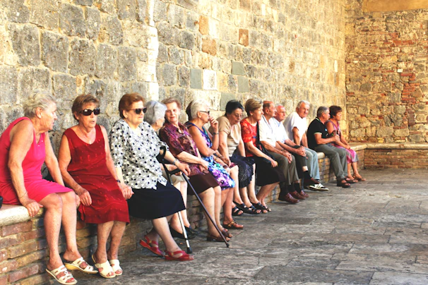A group of elderly people are seated along a stone wall in a historic setting. They are dressed in casual summer clothing and appear to be engaged in conversation. The wall is made of rustic stone bricks, contributing to an old-world charm.