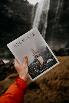 A hand clad in an orange jacket holds up a magazine titled 'Rucksack', featuring an image of a sailing ship on a stormy sea. The background displays a large waterfall cascading down a rugged landscape.