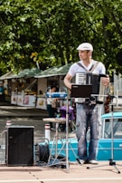 A street musician plays an accordion while standing on a wooden platform. He wears a white shirt and a cap and is surrounded by musical equipment including a laptop, speakers, and cables. In the background, market stalls and trees can be seen, indicating a lively outdoor setting.