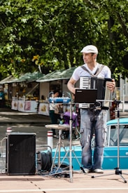 A street musician plays an accordion while standing on a wooden platform. He wears a white shirt and a cap and is surrounded by musical equipment including a laptop, speakers, and cables. In the background, market stalls and trees can be seen, indicating a lively outdoor setting.