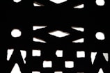 A pattern of geometric shapes, including circles and triangles, is created by light filtering through a perforated surface. The contrast between the illuminated background and the dark structure forms a striking design.