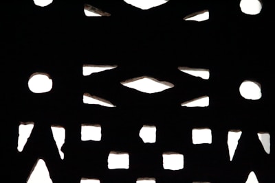 A pattern of geometric shapes, including circles and triangles, is created by light filtering through a perforated surface. The contrast between the illuminated background and the dark structure forms a striking design.