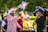 A man wearing a suit covered in British flag patterns, along with a matching hat and sunglasses, waves a flag featuring similar designs. He is smiling broadly towards a cameraman who is filming him. Several people are around, including a woman in a yellow shirt and white hat. They are outdoors with a green, park-like background.