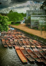 A collection of wooden boats is lined up on a serene river, with lush greenery and a modern building in the background. The sky is overcast, casting a moody atmosphere over the scene.