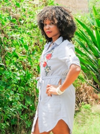 A woman with curly hair stands confidently outdoors, surrounded by lush green foliage. She is wearing a striped dress with floral embroidery, featuring red roses and white flowers. Her hands rest on her hips, and she wears a white beaded bracelet.