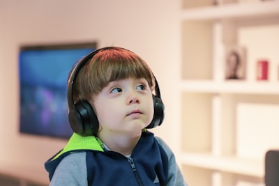 A young child wearing large black headphones, gazing upward with a thoughtful expression. The child is dressed in a blue and lime green hoodie. In the blurred background, there is a television and some blurred shelves with books and photo frames.