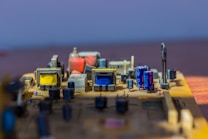 A close-up of an electronic circuit board featuring various components like capacitors, transformers, and other electronic parts. The board has a mix of blue, yellow, and orange components arranged neatly on a flat surface, illuminated by bright light.