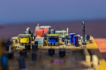 A close-up of an electronic circuit board featuring various components like capacitors, transformers, and other electronic parts. The board has a mix of blue, yellow, and orange components arranged neatly on a flat surface, illuminated by bright light.