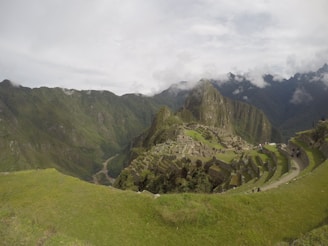 An ancient stone city with terraced fields set against a backdrop of lush, green mountains. Mist and clouds drift around the peaks, adding a sense of mystery. The landscape is expansive, showing a blend of architectural marvel and natural beauty.