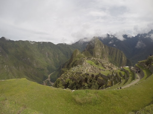 An ancient stone city with terraced fields set against a backdrop of lush, green mountains. Mist and clouds drift around the peaks, adding a sense of mystery. The landscape is expansive, showing a blend of architectural marvel and natural beauty.