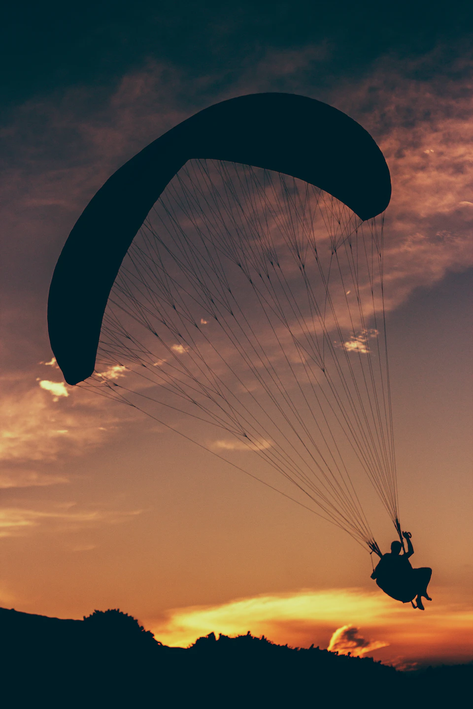 Person suspended in mid-air, silhouetted against orange sky with clouds and flying paraglider