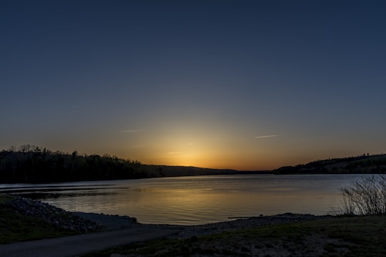 A serene landscape with a calm lake reflecting the colors of the sky, surrounded by silhouettes of trees and gentle hills under a gradient blue to orange sunset.
