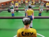 A close-up view of a foosball table game with focus on a yellow-shirted foosball figurine in the foreground. The figurine has the name 'Eddy' written on its back. Other figurines in green and white shirts are visible in the background, lined up on metal rods. The table has a bright green surface with white markings.