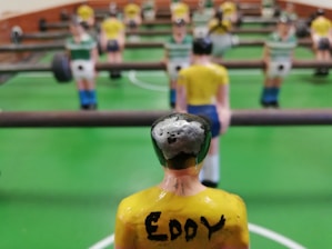A close-up view of a foosball table game with focus on a yellow-shirted foosball figurine in the foreground. The figurine has the name 'Eddy' written on its back. Other figurines in green and white shirts are visible in the background, lined up on metal rods. The table has a bright green surface with white markings.