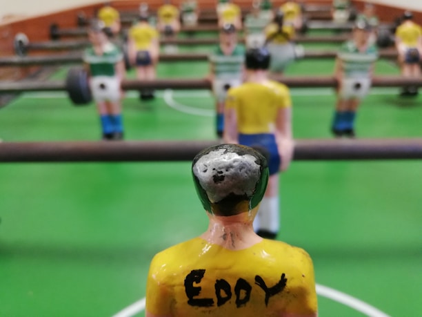 A close-up view of a foosball table game with focus on a yellow-shirted foosball figurine in the foreground. The figurine has the name 'Eddy' written on its back. Other figurines in green and white shirts are visible in the background, lined up on metal rods. The table has a bright green surface with white markings.