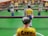 A close-up view of a foosball table game with focus on a yellow-shirted foosball figurine in the foreground. The figurine has the name 'Eddy' written on its back. Other figurines in green and white shirts are visible in the background, lined up on metal rods. The table has a bright green surface with white markings.