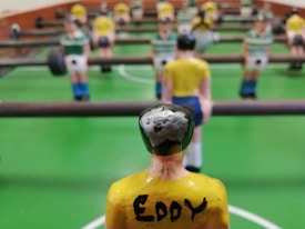 A close-up view of a foosball table game with focus on a yellow-shirted foosball figurine in the foreground. The figurine has the name 'Eddy' written on its back. Other figurines in green and white shirts are visible in the background, lined up on metal rods. The table has a bright green surface with white markings.