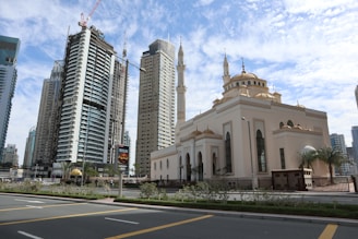 A cityscape featuring a mosque with traditional Islamic architecture, recognizable by its domes and minarets, situated alongside modern high-rise buildings. The sky is partially cloudy, and the streets are lined with greenery and streetlamps.