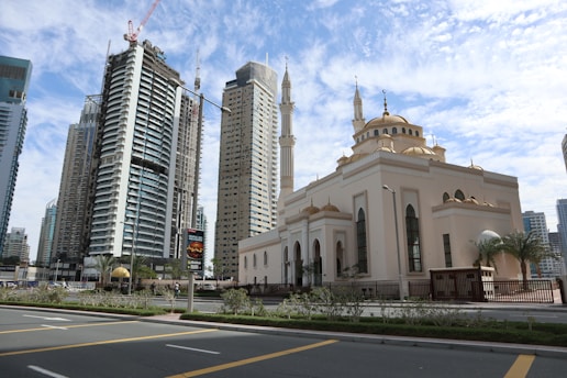 A cityscape featuring a mosque with traditional Islamic architecture, recognizable by its domes and minarets, situated alongside modern high-rise buildings. The sky is partially cloudy, and the streets are lined with greenery and streetlamps.