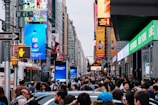 A crowded urban street filled with people and vehicles. Large illuminated advertisements and billboards line the buildings, displaying bright and colorful images and texts. Traffic signals and street signs are present, and a city bus is visible in the midst of the crowd.