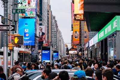 A crowded urban street filled with people and vehicles. Large illuminated advertisements and billboards line the buildings, displaying bright and colorful images and texts. Traffic signals and street signs are present, and a city bus is visible in the midst of the crowd.