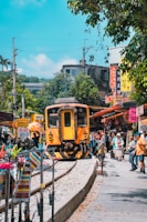A vibrant street scene with a yellow train passing through a market area. The area is bustling with people walking, shopping, and observing. Colorful stalls line both sides of the tracks, displaying various products and signs with Chinese characters. Trees and lush greenery are visible in the background, and the sky is clear and blue.
