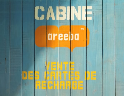 A wooden wall painted in blue with text printed on it. The text includes the word 'CABINE' in white, an orange speech bubble with the word 'areeba' inside, and 'VENTE DES CARTES DE RECHARGE' in yellow below.