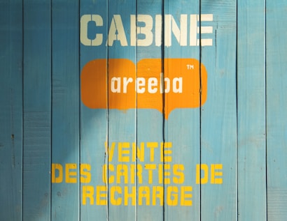 A wooden wall painted in blue with text printed on it. The text includes the word 'CABINE' in white, an orange speech bubble with the word 'areeba' inside, and 'VENTE DES CARTES DE RECHARGE' in yellow below.