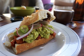 A sandwich with a crispy baguette is filled with guacamole, sliced jalapenos, and onions, served on a white plate. In the background, there's a cup of coffee and a bowl, with blurred figures sitting at a wooden table.