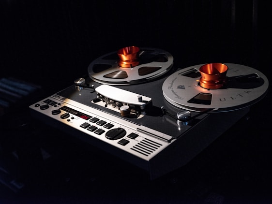 A vintage reel-to-reel tape recorder with large metallic spools and orange hubs is positioned against a dark background. The device features an intricate array of buttons and knobs on its control panel, with a digital display showing '005.' The lighting emphasizes the shiny metallic surface of the reels, creating a nostalgic and classic ambiance.