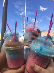Several hands are holding colorful slushie drinks with domed lids and pink straws. The slushies display vibrant hues, including pink and blue, against a backdrop of a bright blue sky with a few clouds. A ferris wheel and a pole are visible in the background, suggesting a setting of an amusement park or fair.
