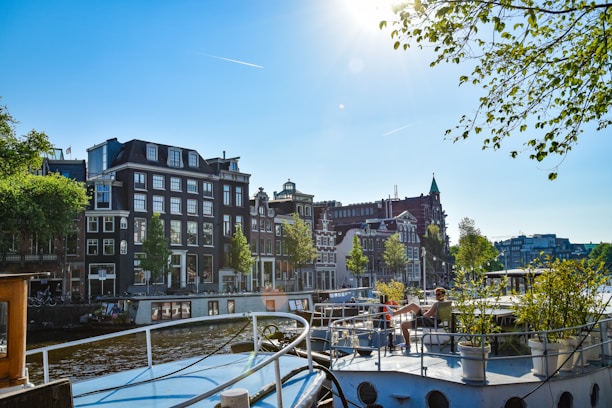 A scenic canal view featuring traditional Dutch buildings with gabled roofs along one side. The canal has boats moored, and a person sits leisurely on the deck of one boat. Trees line both sides, and the sun shines brightly in the clear blue sky, enhancing the vibrant atmosphere.