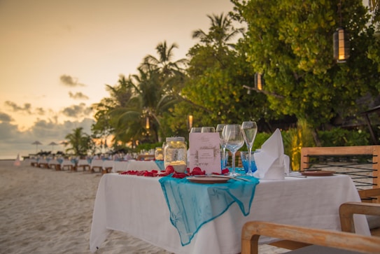 A table set for an outdoor dinner on a sandy beach near the ocean, with lush green palm trees in the background. The table is adorned with red and turquoise decorations, wine glasses, and a menu card, creating a romantic and inviting atmosphere as the sun sets.