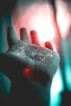 A hand is extended with shimmering glitter resting on its surface, illuminated by a soft, colorful light in the background creating a magical atmosphere.
