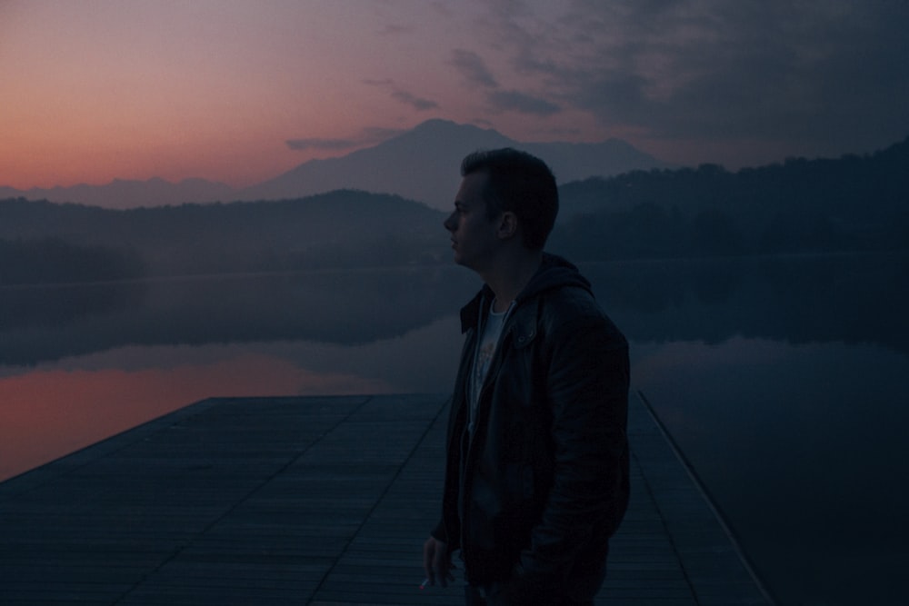 man in black hoodie standing on dock near body of water during golden hour