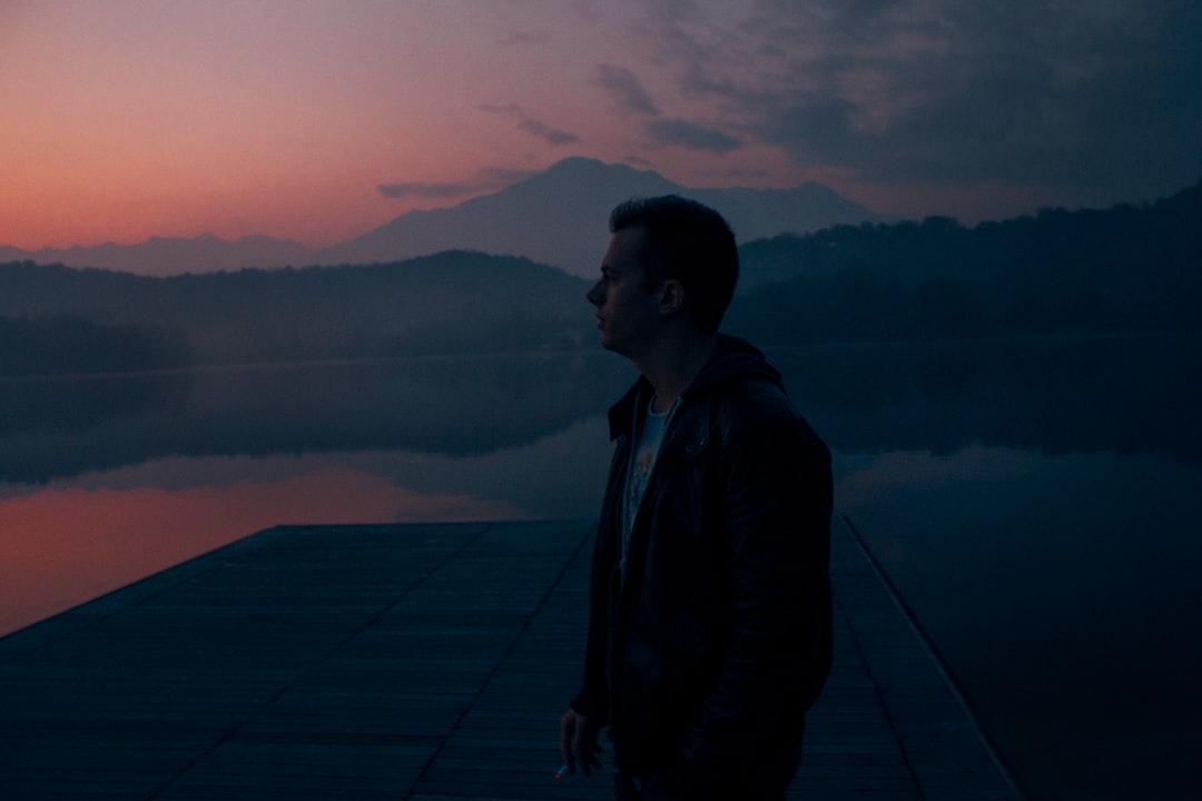 man in black hoodie standing on dock near body of water during golden hour