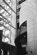 A modern architectural space with geometric shapes, featuring a spiral staircase encased in glass and metal. The building's exterior is adorned with large, reflective windows that accentuate the sleek, contemporary design.