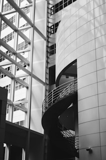 A modern architectural space with geometric shapes, featuring a spiral staircase encased in glass and metal. The building's exterior is adorned with large, reflective windows that accentuate the sleek, contemporary design.